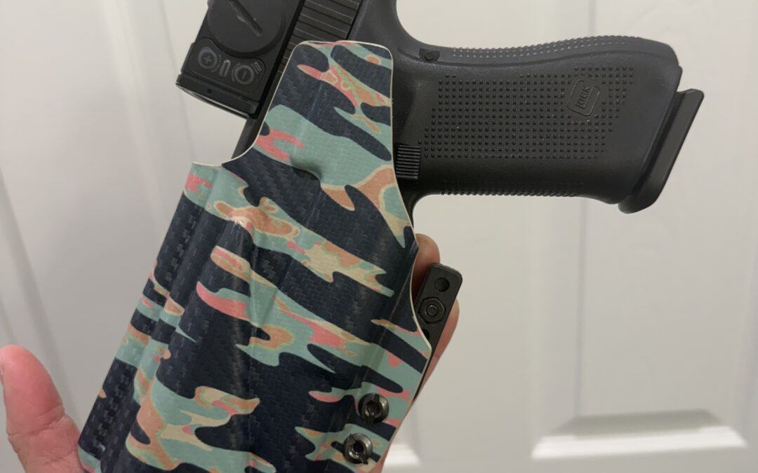 Black handgun secured in a camouflage tactical holster, held in a hand against a light background.