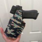 Black handgun secured in a camouflage tactical holster, held in a hand against a light background.