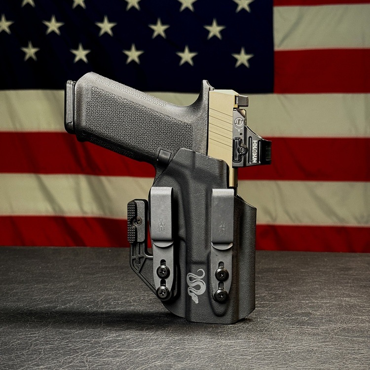 Black holstered semi-automatic handgun with a tan slide on a dark surface, with the American flag in the background.