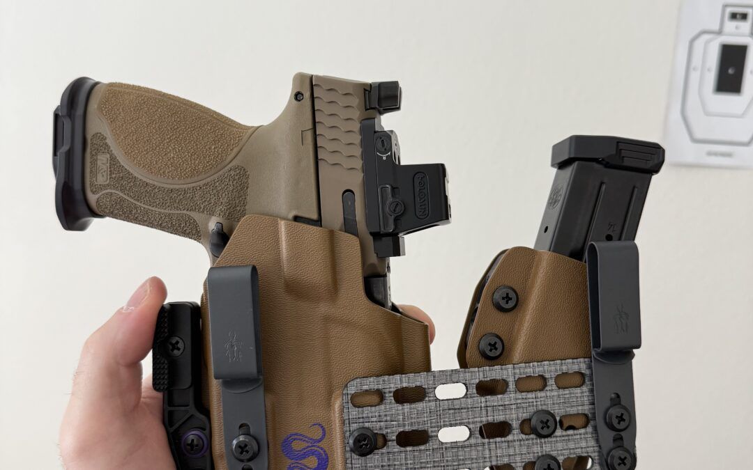 Close-up of a hand holding a tan rifle lower receiver with a textured grip and mounted magazine/rail system visible in the foreground.