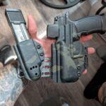 Person wearing camouflage sleeve holds a pistol in a holster with a detachable magazine nearby on a wooden floor surface in the background.